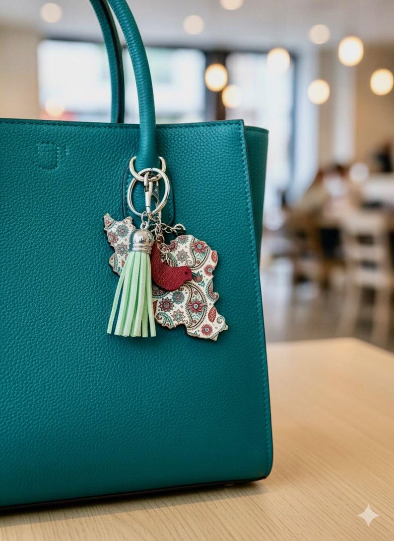 May include: Teal leather handbag with a decorative keychain. The keychain features a light green tassel, a red bird, and a wooden cutout in the shape of a country with a paisley design. The bag has a textured surface.