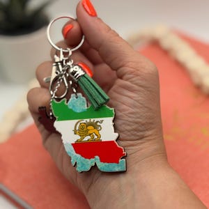 May include: A keyring featuring a map-shaped pendant of Iran, coloured with green, white, and red horizontal stripes and a central gold lion and sun emblem. The pendant includes a small green suede tassel and a silver-toned metal ring and clasp.