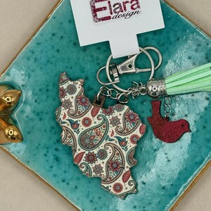 May include: A keychain featuring a wooden charm in the shape of a country, adorned with a paisley pattern in red, blue, and cream. It includes a red bird charm and a mint green tassel. The tag reads "Elara design".
