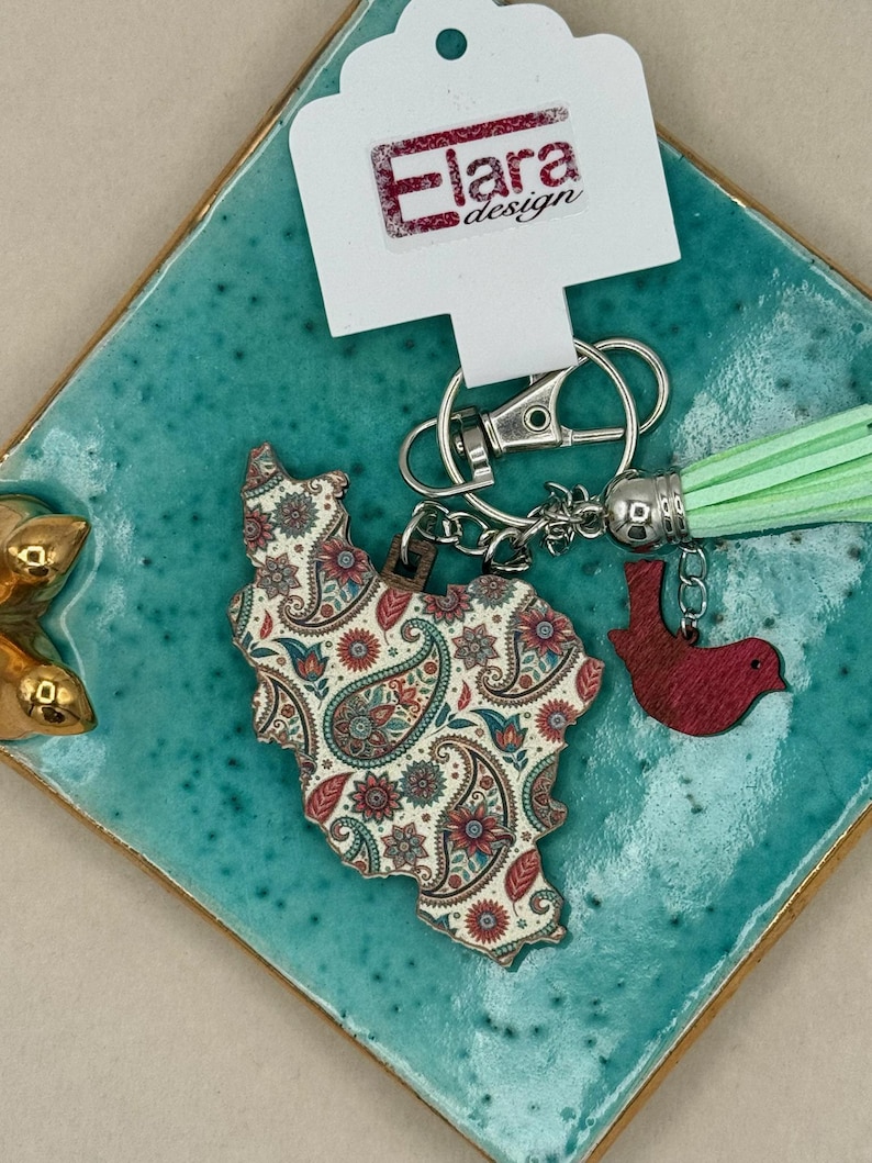 May include: A keychain featuring a wooden charm in the shape of a country, adorned with a paisley pattern in red, blue, and cream. It includes a red bird charm and a mint green tassel. The tag reads "Elara design".