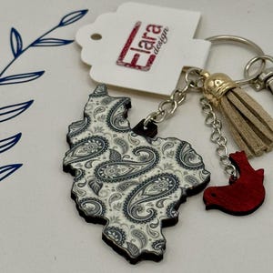 May include: A wooden keychain featuring a paisley design in white and gray. The keychain has a small red bird charm and a tan tassel. The tag reads "Flara design".