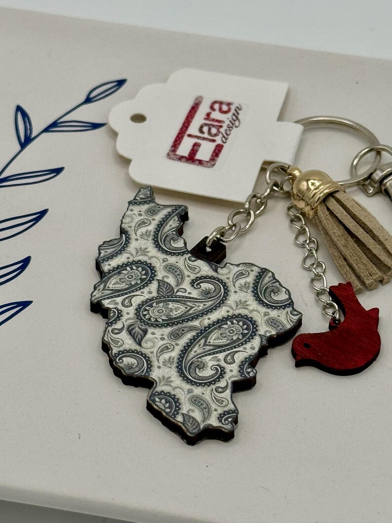 May include: A wooden keychain featuring a paisley design in white and gray. The keychain has a small red bird charm and a tan tassel. The tag reads "Flara design".