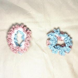May include: Two handmade crochet scrunchies. One is pink, white, and blue, and the other is blue and pink. The scrunchies are circular and have a ruffled edge. They are made of soft yarn and are suitable for various hairstyles.