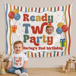 May include: A white banner with the words "Ready Two Party" in colorful letters, balloons, and a baby's face. The banner is for Harley's 2nd birthday. A baby wearing a matching shirt and jeans sits in front of the banner.