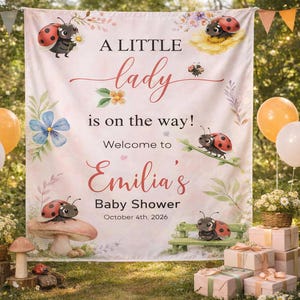 May include: A baby shower banner with the text "A LITTLE lady is on the way! Welcome to Emilia's Baby Shower October 4th, 2026." The banner features ladybug illustrations and floral accents in a soft pink and green color scheme.