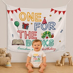 May include: A white fabric backdrop and a matching white baby bodysuit featuring the text ONE FOR THE BOOKS and Camden's birthday. The design includes a green caterpillar, a red party hat, a gift box, a green balloon, and an open book illustration.