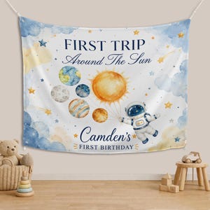 May include: A white fabric backdrop featuring a watercolor space theme with blue clouds, yellow stars, and planets. The text reads FIRST TRIP Around The Sun and Camden's FIRST BIRTHDAY. An astronaut holds strings attached to a large yellow sun.