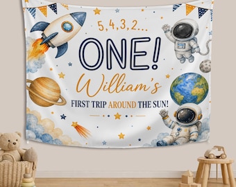 May include: A white fabric wall hanging featuring a space theme with a rocket, two astronauts, a planet with rings, Earth, and stars. The text reads 5, 4, 3, 2... ONE! William's FIRST TRIP AROUND THE SUN! It includes blue and orange bunting flags.
