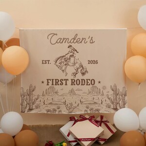 Może przedstawiać: Beżowy, prostokątny baner z tkaniny z brązową ilustracją kowboja na wierzgającym koniu. Tekst głosi "Camden's", "EST. 2026" oraz "FIRST RODEO" nad pustynnym krajobrazem z kaktusami. Białe i beżowe balony otaczają baner.