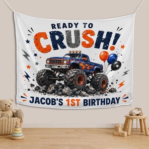 May include: A white fabric wall hanging featuring a blue monster truck with orange flames, surrounded by orange and blue balloons, stars, and lightning bolts. The text reads READY TO CRUSH! and JACOB'S 1ST BIRTHDAY in bold blue and orange letters.