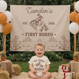 Może przedstawiać: Beżowe tło z tkaniny z brązową ilustracją kowboja na wierzgającym koniu, napisem "Camden's EST. 2026 FIRST RODEO" oraz pustynnym krajobrazem z kaktusami. Niemowlę ma na sobie pasujące beżowe body z napisem "MY FIRST RODEO".