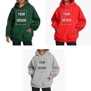 Op de afbeelding: Drie hoodies in verschillende kleuren: bosgroen, rood en grijs. Elke hoodie heeft een voorzak en een capuchon met een trekkoord. De tekst "YOUR DESIGN" staat in een witte rechthoek op de voorkant.