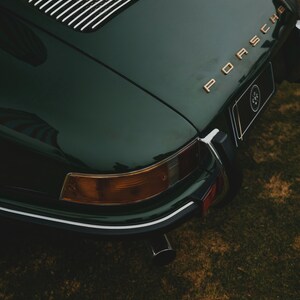 May include: Dark green vintage Porsche car with gold lettering. The rear of the car is visible, showcasing the taillights, exhaust pipe, and the Porsche logo. The car is parked on a grassy surface.
