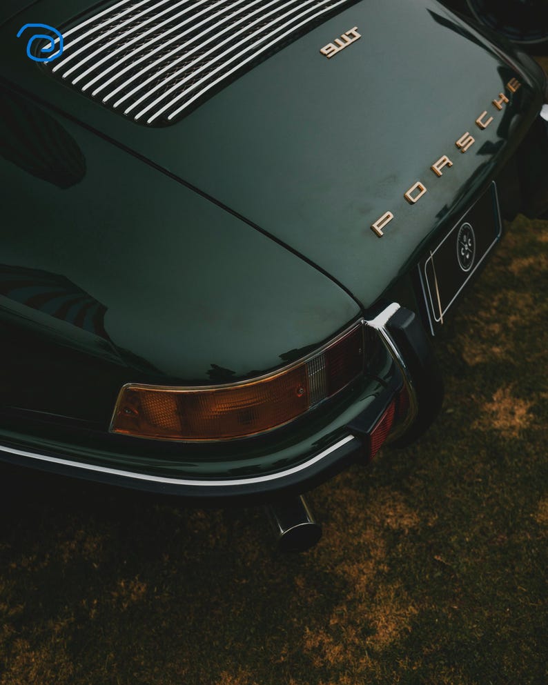 May include: Dark green vintage Porsche car with gold lettering. The rear of the car is visible, showcasing the taillights, exhaust pipe, and the Porsche logo. The car is parked on a grassy surface.
