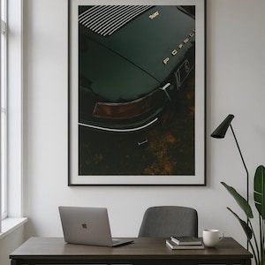 May include: Framed print of a dark green vintage Porsche car. The image shows the rear of the car with gold lettering and a distinctive tail light. The print is displayed above a desk with a laptop, chair, and coffee cup.