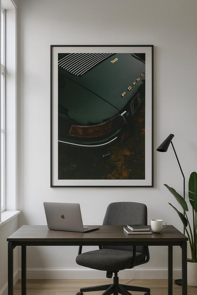 May include: Framed print of a dark green vintage Porsche car. The image shows the rear of the car with gold lettering and a distinctive tail light. The print is displayed above a desk with a laptop, chair, and coffee cup.