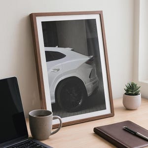May include: A framed print of a white luxury SUV with black wheels, leaning against a wall. The print is in a brown wooden frame. A laptop, coffee mug, notebook, and small plant are on a wooden desk.