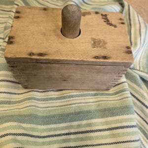 May include: A rectangular wooden butter mold with a light brown finish, featuring a central cylindrical handle on the lid. The box is constructed with visible finger joints at the corners and small metal nails securing the top panel.