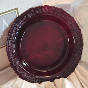 May include: A deep burgundy-colored plate with a decorative, scalloped edge. The plate has an embossed floral and geometric pattern around the rim. The center of the plate has a radial design. The plate is part of a collection.