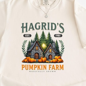 Könnte beinhalten: Cremefarbenes T-Shirt mit dem Text "Hagrid's Pumpkin Farm" und einer Grafik von zwei Steinhäusern, Kürbissen und immergrünen Bäumen. Das Design enthält den Text "Est. 1981" und "Magically Grown".