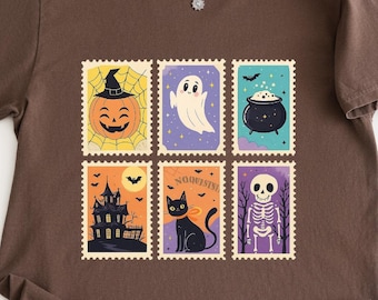 May include: Brown t-shirt featuring six Halloween-themed postage stamp designs. The stamps depict a jack-o'-lantern, ghost, cauldron, haunted house, black cat, and skeleton. A silver necklace with a pendant is visible.