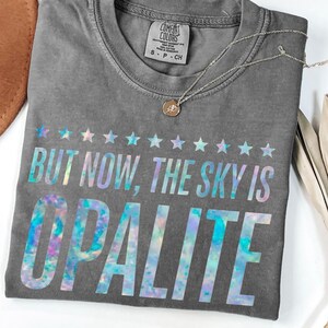 Pode incluir: Uma camiseta cinza Comfort Colors com o texto "BUT NOW, THE SKY IS OPALITE" em uma fonte holográfica. A camiseta tem gola redonda e mangas curtas. Um colar de prata com um pequeno pingente está sobre a camiseta.