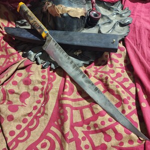 Könnte beinhalten: Ein langes, handgefertigtes Messer mit einem Holzgriff mit gelben Akzenten. Die Klinge ist silberfarben mit einer strukturierten Oberfläche. Das Messer liegt auf einem gemusterten rot-goldenen Stoff, mit einer schwarzen Scheide und anderen Objekten im Hintergrund.