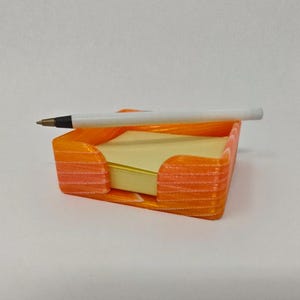 Könnte beinhalten: Ein orangefarbener, 3D-gedruckter Haftnotizhalter mit einem Stapel gelber Haftnotizen und einem weißen Stift. Der Halter hat eine rechteckige Form mit gebogenen Seiten und steht auf einer weißen Oberfläche.