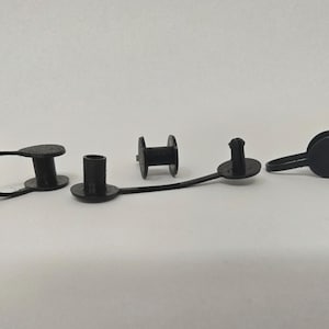 Puede incluir: Cuatro piezas de plástico negro están dispuestas sobre una superficie blanca. Las piezas incluyen dos con un lazo, una con forma de carrete y otra con forma cilíndrica. Las piezas parecen pequeñas y de plástico mate.