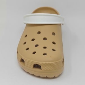 May include: A tan slip-on clog featuring a white pivoting heel strap. The shoe has a rounded toe, a textured border along the base, and multiple circular ventilation holes across the top surface, providing a functional and simple design.