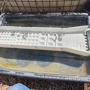 Peut inclure: Un bac de lavage d'or en plastique noir avec un plateau à riffles en plastique blanc. Le plateau présente différents motifs pour séparer les matériaux. Le bac est rempli d'eau et est à l'extérieur.
