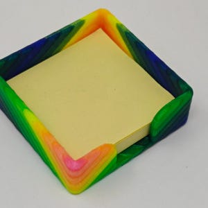 Könnte beinhalten: Ein quadratischer Schreibtisch-Organizer mit einem Regenbogen-Farbverlauf an den Kanten, der einen Stapel gelber Haftnotizen enthält. Die Farben gehen von Rosa über Orange, Grün bis Blau.