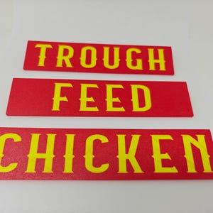Puede incluir: Tres letreros rectangulares rojos con letras amarillas. El letrero superior dice "TROUGH", el letrero del medio dice "FEED" y el letrero inferior dice "CHICKEN". Estos letreros son probablemente para una granja o agricultura.