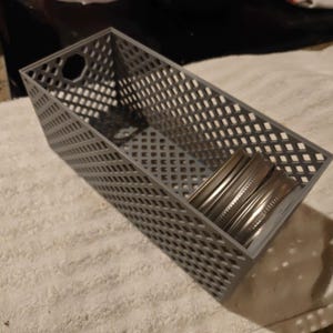 May include: A rectangular gray storage bin with a diamond-shaped lattice pattern on the sides and base. The bin features a circular cutout handle on each end and contains two circular metallic jar lids resting inside the container.