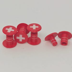 Könnte beinhalten: Eine Ansammlung von fünf roten zylindrischen Verschlüssen mit einem weißen Kreuzsymbol auf der Oberseite. Drei der Verschlüsse stehen aufrecht, während zwei auf der Seite liegen und das hohle Innere sowie weiße Ringdetails zeigen.