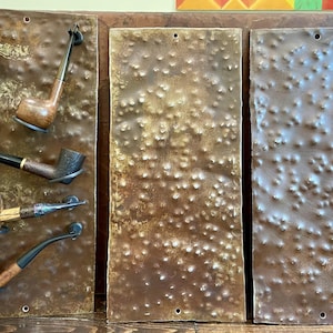 May include: A display of five tobacco pipes mounted on a textured, brown metal panel. The pipes vary in shape and color, with wooden bowls and black stems. The panel has a rustic, industrial aesthetic.