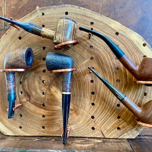 May include: An assortment of wooden tobacco pipes with various shapes and finishes. The pipes are displayed on a wooden plaque with a natural edge and a series of drilled holes. The pipes have copper-colored holders.