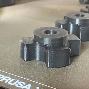 May include: A dark gray, 3D-printed knob with a circular top and four curved extensions. The knob has a hole in the center and is sitting on a textured surface. Another similar knob is visible in the background.