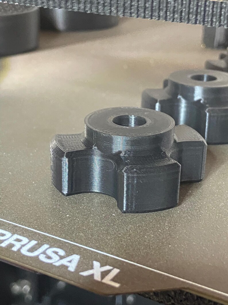 May include: A dark gray, 3D-printed knob with a circular top and four curved extensions. The knob has a hole in the center and is sitting on a textured surface. Another similar knob is visible in the background.