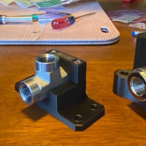 May include: Three black metal mechanical components with silver fittings are displayed on a wooden surface. The components vary in shape, including a cube, an L-shaped piece, and a piece with a circular base. Tools are visible in the background.