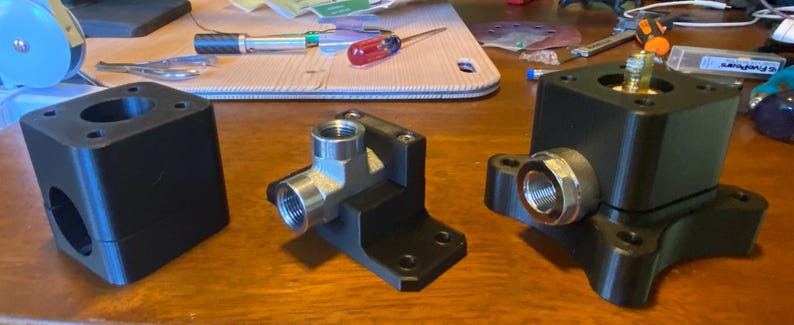 May include: Three black metal mechanical components with silver fittings are displayed on a wooden surface. The components vary in shape, including a cube, an L-shaped piece, and a piece with a circular base. Tools are visible in the background.