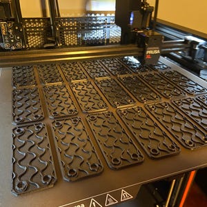 May include: A 3D printer bed with multiple black, rectangular parts with a wavy pattern and rounded corners. The parts are arranged in rows on the printer bed. The printer has the Prusa XL logo.