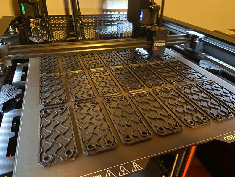May include: A 3D printer bed with multiple black, rectangular parts with a wavy pattern and rounded corners. The parts are arranged in rows on the printer bed. The printer has the Prusa XL logo.