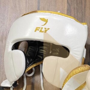 May include: White boxing headgear with gold trim and the word "FLY" in gold lettering. The headgear has a black interior and a chin strap. A gold bird graphic is above the word "FLY".