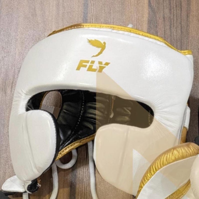May include: White boxing headgear with gold trim and the word "FLY" in gold lettering. The headgear has a black interior and a chin strap. A gold bird graphic is above the word "FLY".