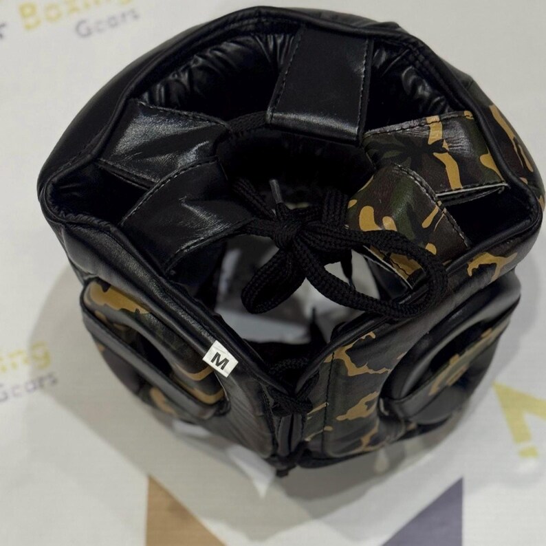 Puede incluir: Casco de boxeo negro y camuflaje con cierre de cordones. El casco tiene un interior acolchado y una etiqueta marcada con la letra "M". El patr&oacute;n de camuflaje incluye tonos de verde, marr&oacute;n y beige.