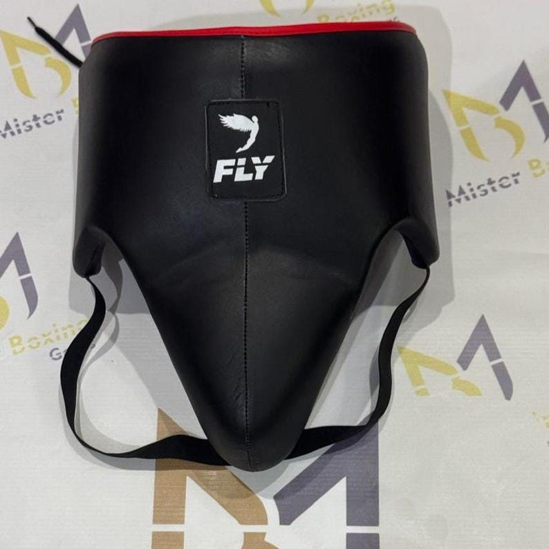 May include: Black protective cup with a red trim and the word "FLY" printed on the front. The cup has a white bird logo and black straps for securing. This item is designed for sports and martial arts.
