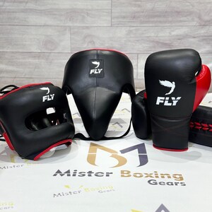 May include: A set of black and red boxing gear, including a head guard, face protector, and gloves. The gloves and headgear have the word "FLY" and a white bird logo. The gear is displayed on a white surface.