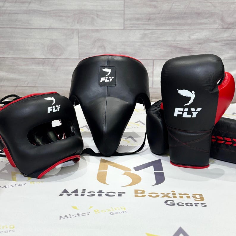 May include: A set of black and red boxing gear, including a head guard, face protector, and gloves. The gloves and headgear have the word "FLY" and a white bird logo. The gear is displayed on a white surface.