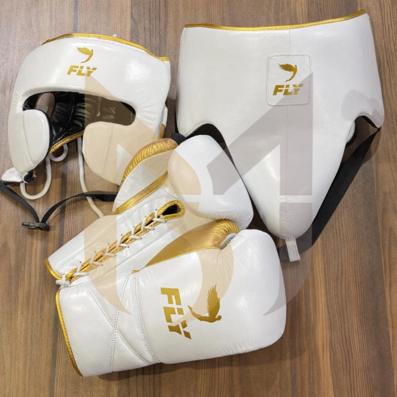 May include: A white boxing headguard, gloves, and groin protector set with gold detailing. The gloves are laced up the centre. The brand name "FLY" and a bird logo are printed on the items. The set is displayed on a wooden surface.